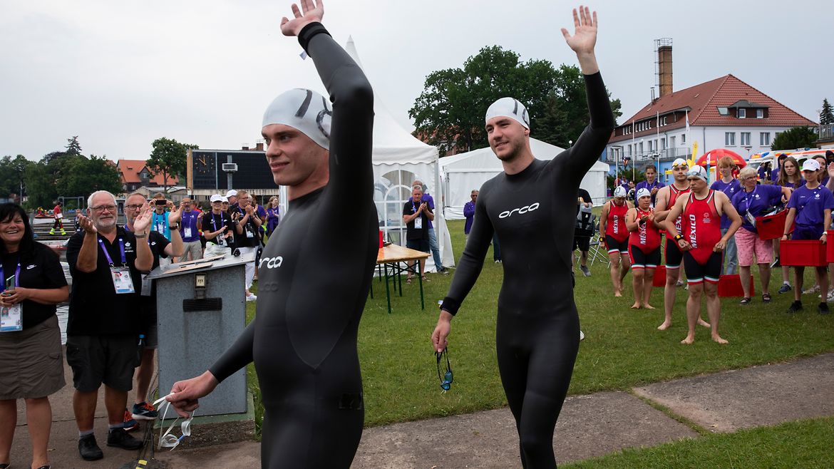 Unified-Schwimmer Leo Heckel und Adrian Schlüter in Neopren-Anzügen winken in die Kamera.