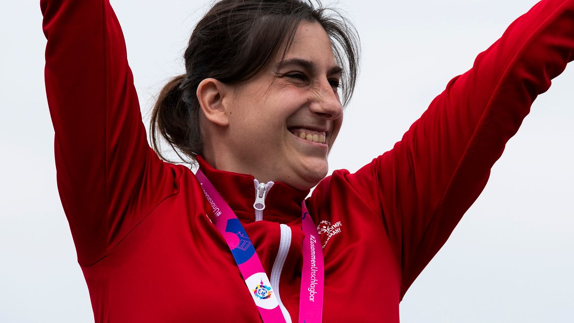 Patrizia Spaulding mit Goldmedaille in roter Trainingsjacke reißt beide Arme hoch und lacht.