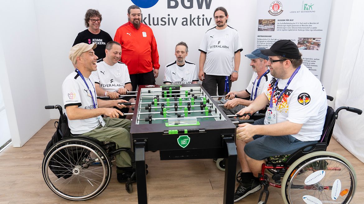 Vier Männer im Rollstuhl spielen an einem Tischfußball-Tisch. Im Hintergrund 4 weitere Männer in Trikots und eine Infotafel "Inklusives Kickern beim FC St. Pauli". An der Wand daneben der Schriftzug "BGW inklusiv aktiv"