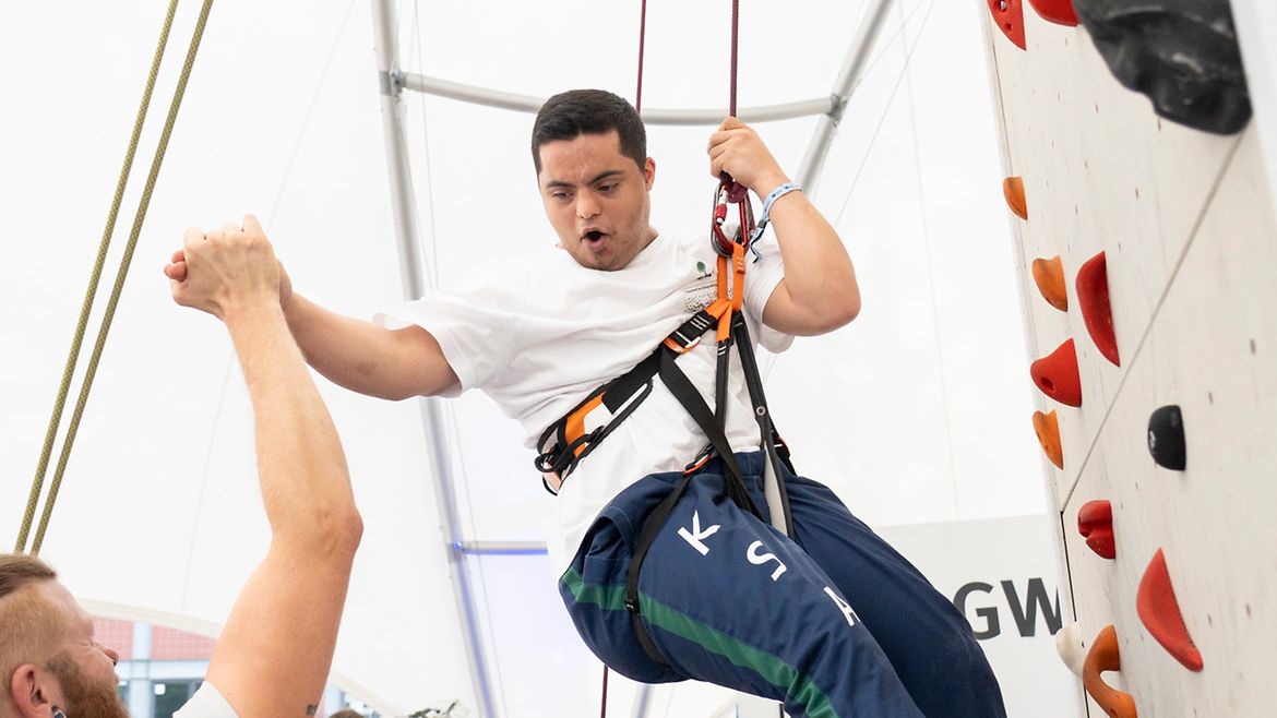 Ein junger Mann wird am Kletterturm abgeseilt. Eine Frau sichert ihn, ein Mann klatscht mit ihm ab.