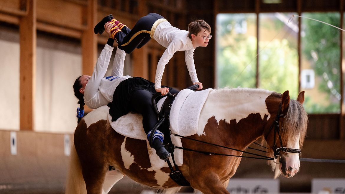 Ein Mädchen liegt auf einem Pferd und hält dabei über sich einen Jungen an den Füßen hoch. Er stützt sich mit den Händen am Sattel ab.