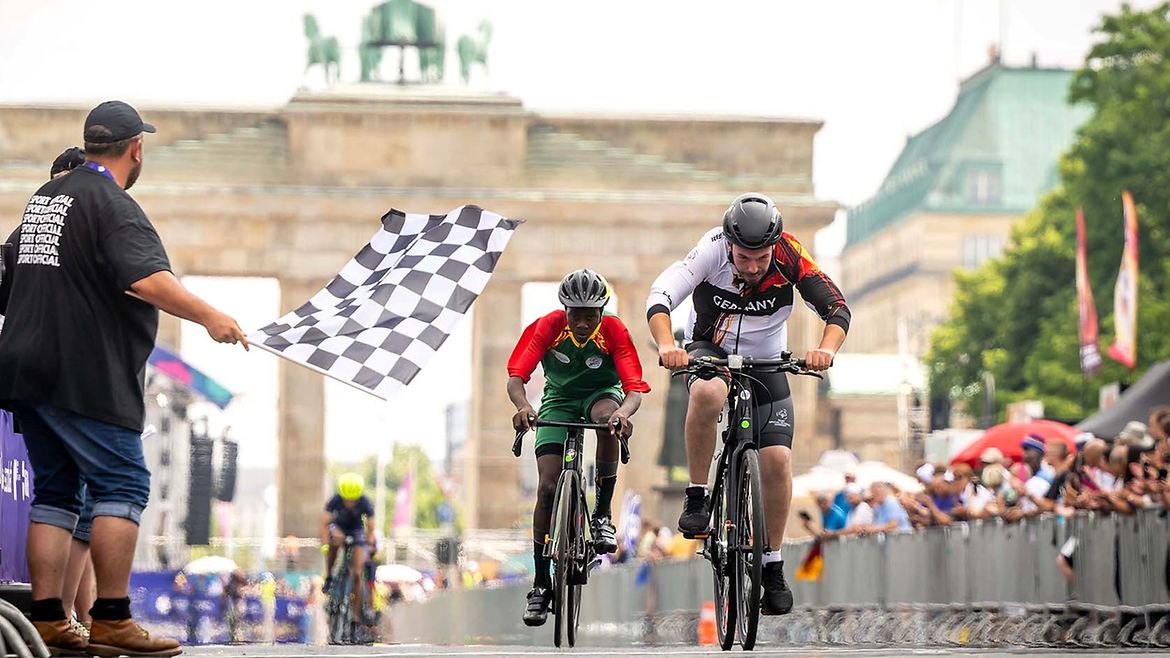 Zwei Radrennfahrer auf der Rennstrecke. Links ein Mann mit Zielflagge, rechts Zuschauende am Rand der Straße. Im Hintergrund das Brandenburger Tor.