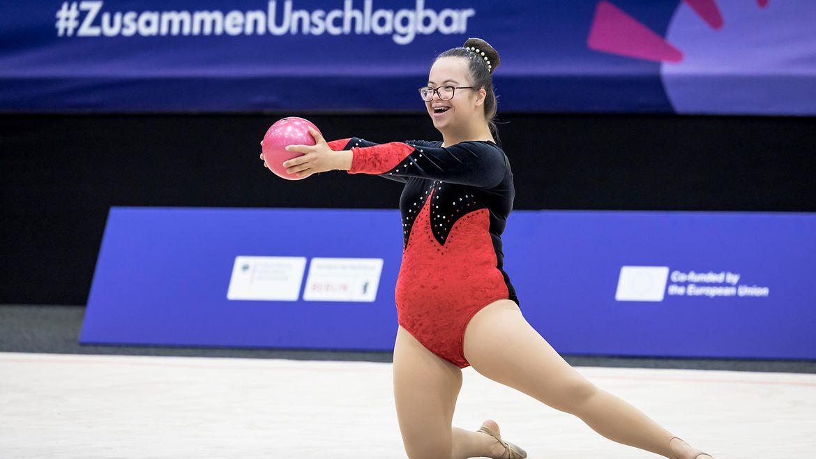 Ivana-Helena Duric hält mit ausgestreckten Armen und beiden Händen anmutig einen Ball vor sich, ein Bein ausgestreckt, eins kniend. Im Hintergrund der Schriftzug #ZusammenUnschlagbar