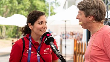 Mehtap Özgül steht neben Ralf Podszus, der ihr ein Mikrophon an den Mund hält. Im Hintergrund der BGW-Stand.