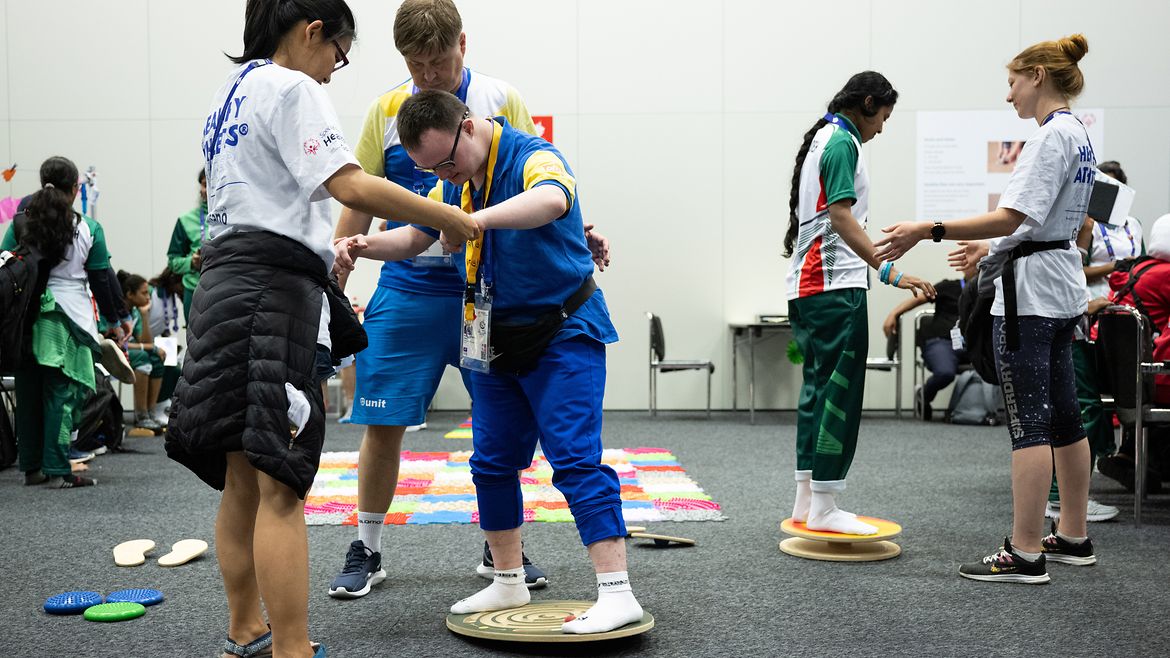 ein Athlet und eine Athletin stehen jeweils auf einem Balanceboard, um sie herum Helfende.