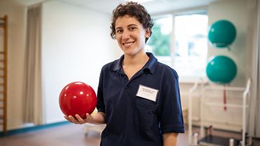 Eine Physiotherapeutin steht in einem Reha-Raum und hält einen kleinen Medizinball in der Hand