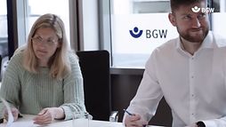 Zwei Beschäftigte der BGW sitzen bei einem Arbeitstreffen an einem Tisch. Auf dem Fenster hinter ihnen klebt ein BGW-Logo