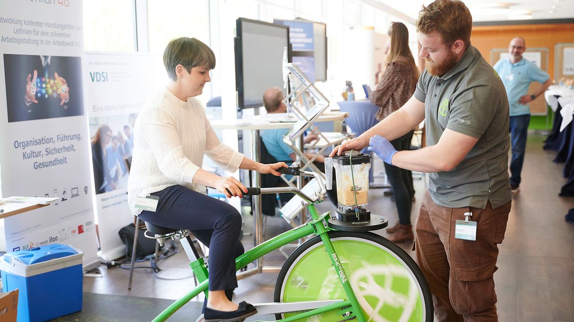 Eine Teilnehmerin der BGW Sifa-Fachtagung 2023 sitzt auf einem Trainings-Fahrrad, mit dem offenbar ein Mixer betrieben wird, der über dem Vorderreiffen angebracht ist.