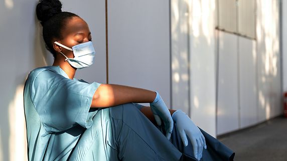 Pflegerin mit Mundschutz und hochgebundenen Haaren sitzt in einem Gang, die Augen geschlossen, und wirkt erschöpft. Schräg stehende Sommersonne beleuchtet Teile von Arm und Gesicht