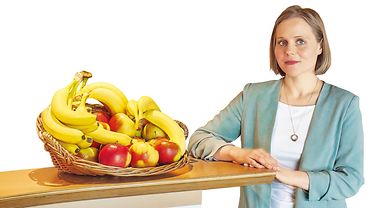 Lena Gombert steht an einem Tisch, auf dem ein großer Obstkorb mit Äpfeln und Bananen steht