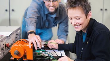 Eine Frau sitzt an einem Arbeitstisch mit einem technischen Gerät mit vielen Kabeln - ein Mann steht neben ihr und zeigt ihr etwas an dem Gerät.