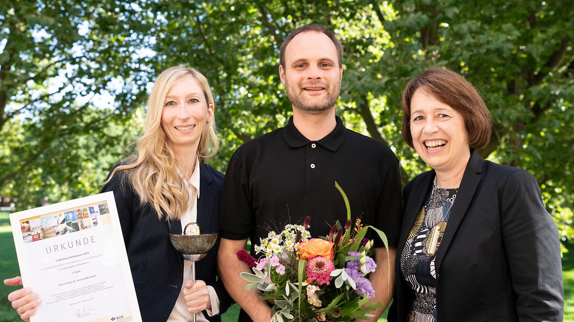 Claudia Drechsel-Schlund posiert lächelnd mit Nicole Schröder und Simon Gaußmann, die den Siegerpokal, eine Urkunde und Blumen in den Händen halten.