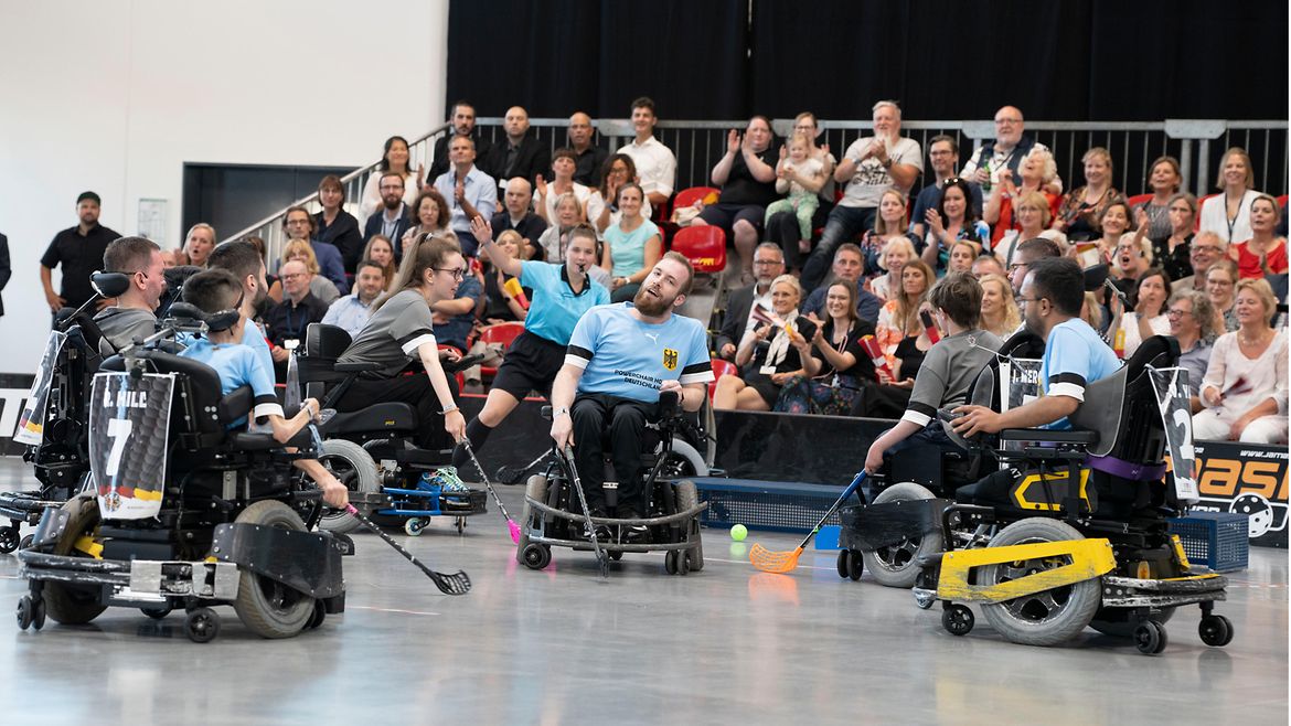 Hockey-Spielerinnen und -Spieler in elektrischen Rollstühlen in einer Sporthalle, im Hintergrund applaudiert das Publikum.
