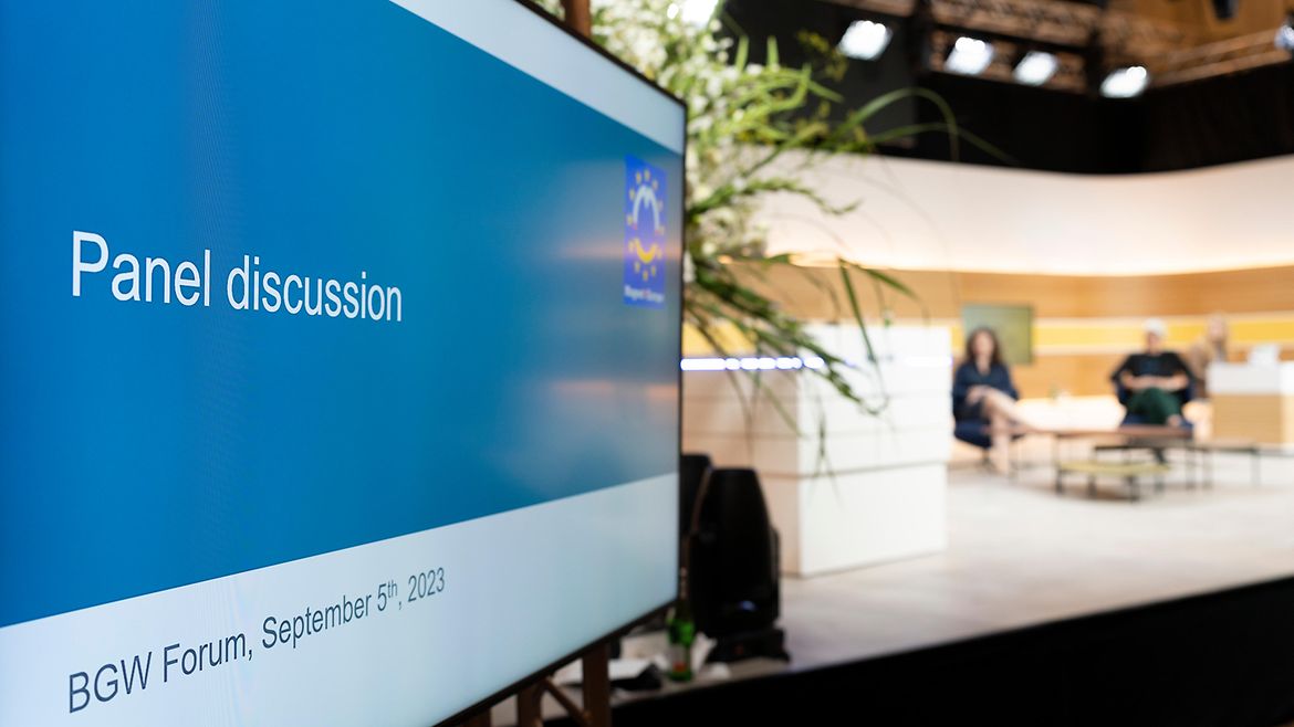 Auf einem Bildschirm im Vordergrund steht "Panel discussion", im unscharfen Hintergrund sitzen 2 Menschen auf einer beleuchteten Bühne