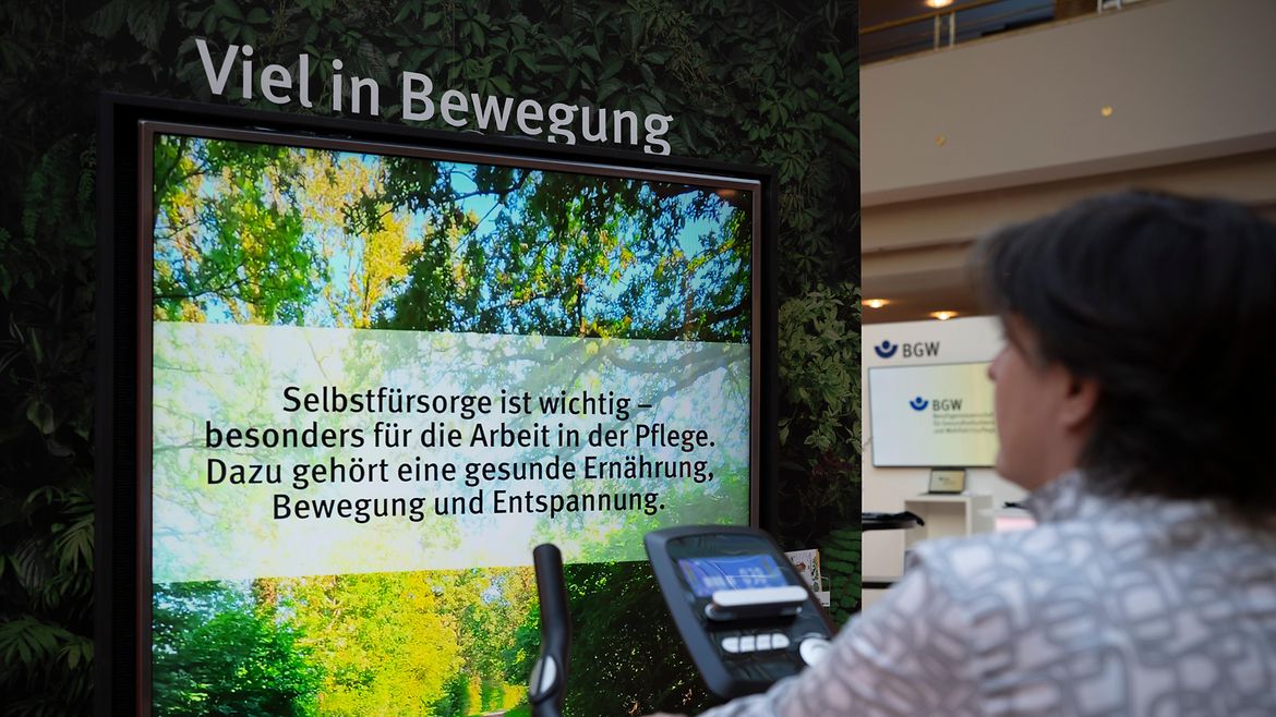 Eine Frau sitzt auf einem Fahrradergometer vor einem Bildschirm, auf dem steht "Viel in Bewegung: Seobstfürsorge ist wichtig - besonders für die Arbeit in der Pflege. Dazu gehört eine gesunde Ernährung, Bewegung und Entspannung." 