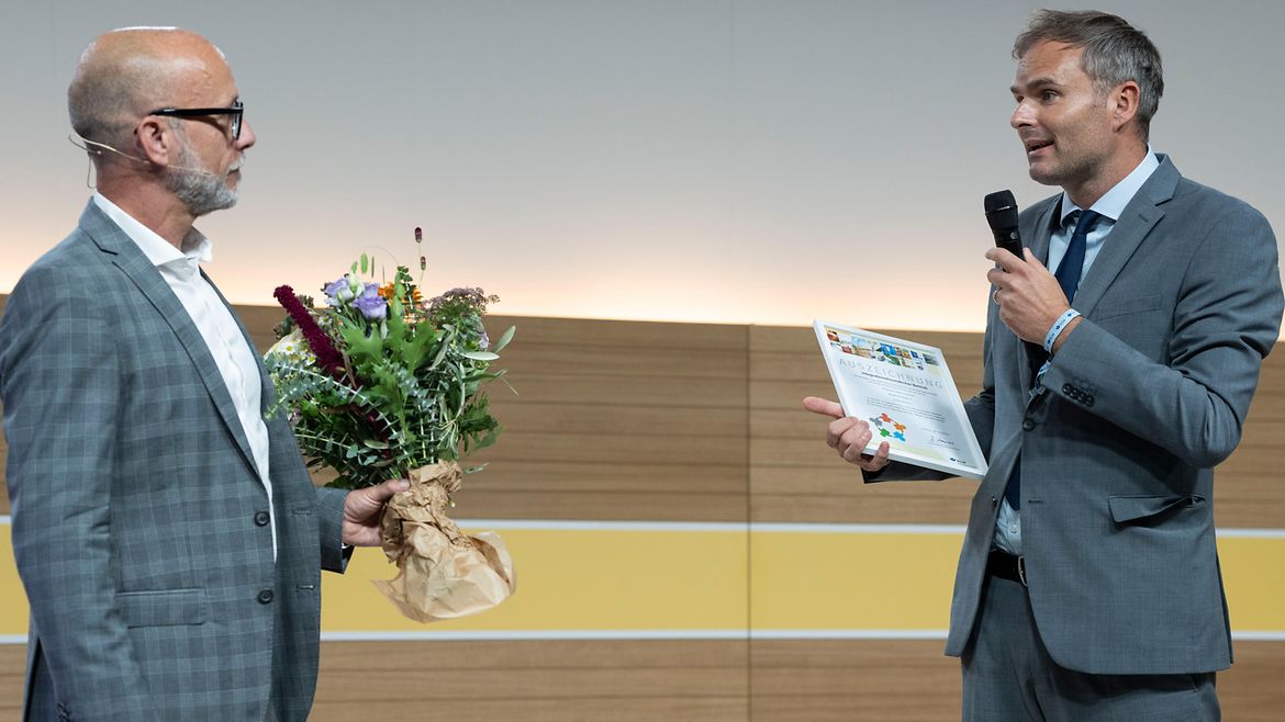 Zwei Männer in Anzug stehen einander zugewwandt auf einer Bühne, der Linke hat einen Blumenstrauß in der Hand, der rechte spricht in ein Mikrofon und hat eine Urkunde in der Hand