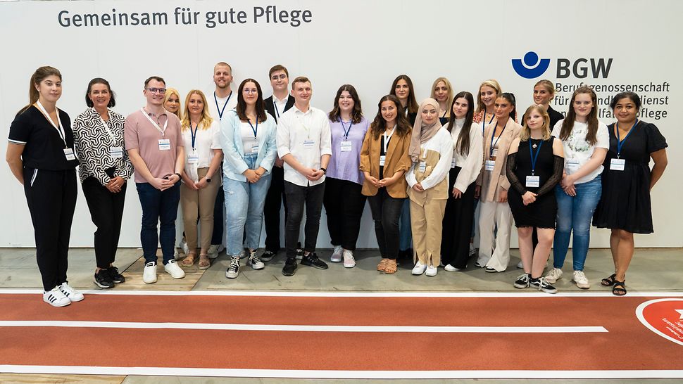 19 junge und 2 erwachsene Personen stehen in einer Messehalle vor einer weißen Wand mit BGW-Logo und der Aufschrift "Gemeinsam für gute Pflege".