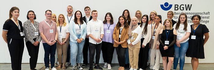 19 junge und 2 erwachsene Personen stehen in einer Messehalle vor einer weißen Wand mit BGW-Logo und der Aufschrift "Gemeinsam für gute Pflege".
