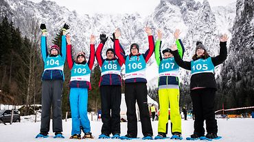 Ein Schneeschuh-Team aus sechs Personen steht in einer Schneelandschaft vor Bergen und streckt die Hände in den Himmel.