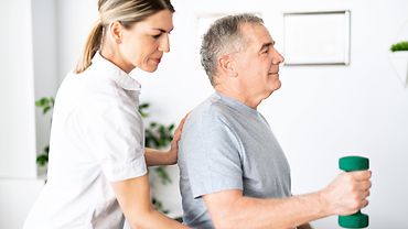 Physiotherapeutin steht hinter einem sitzenden Patienten - sie hat eine Hand an seinem Rücken, die andere an seiner Seite