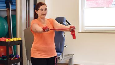 Simone Lorenz bei der Physiotherapie - sie steht und hat die Arme schulterbreit nach vorne ausgestreckt und hält ein Theraband stramm zwischen den Händen. 