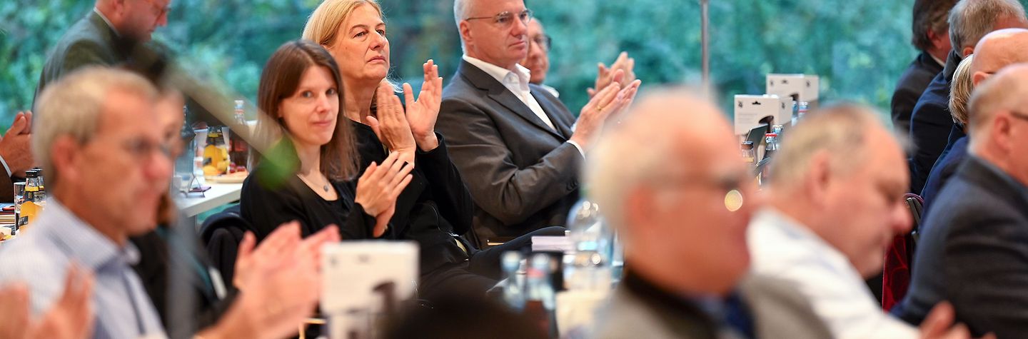 BGW-Vertreterversammlung 2023 in Dresden - Tag 1 Rund zehn Personen applaudieren. Sie sitzen an Tischen und sind von der Seite zu sehen. Einige Personen im Vordergrund sind unscharf abgebildet. Im Hintergrund erblickt man durch große Fenster dichten grünen Baumbewuchs.
