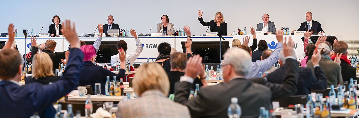 BGW-Vertreterversammlung 2023 in Dresden - Tag 2 Mitglieder der BGW-Verstreterversammlung sitzen in mehreren Reihen an Tischen, auf denen Getränkeflaschen stehen, und stimmen per Handzeichen ab, und man sieht sie von hinten. Im Hintergrund: das Podium mit weiteren sechs Personen, von denen eine die Hand zur Abstimmung hebt.