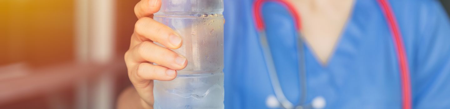 Pflegerin mit Mundschutz und blauem Kassack streckt eine Plastikflasche Wasser nach vorn.