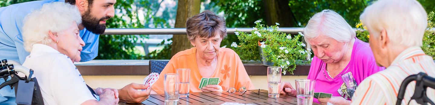 Vier ältere Personen spielen draußen an einem Tisch Karten. Ein Pfleger hält die Spielkarten für eine alte Dame im Rollstuhl. Alle haben ein Glas Wasser vor sich stehen.