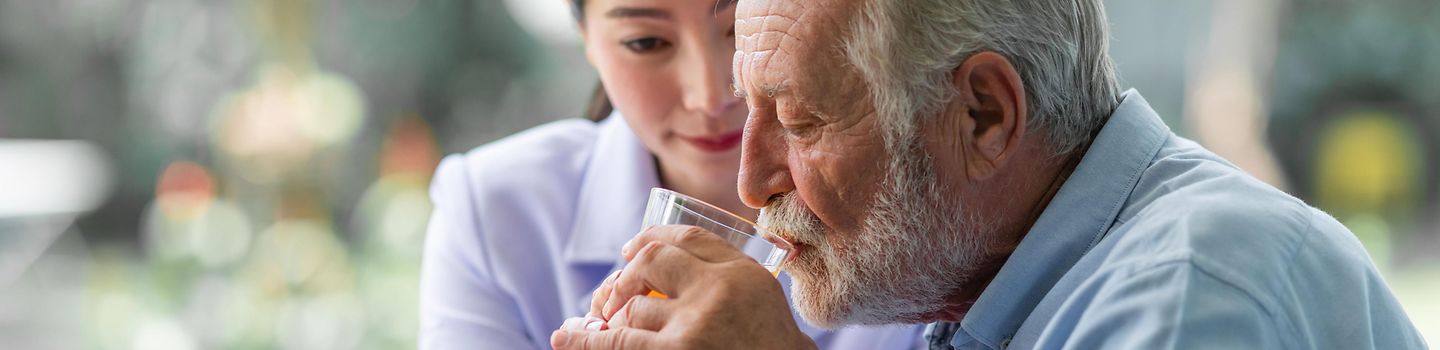 Eine Pflegerin hält einem alten Mann ein Glas mit Saft an den Mund. Beide sitzen in einem Garten.