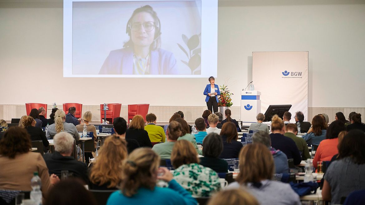 Blick auf die Bühne im Veranstaltungssaal aus dem Publikum, auf der Leinwand ist eine Frau mit Headset zu sehen, die sich anscheinend mit der Moderatorin auf der Bühne unterhält