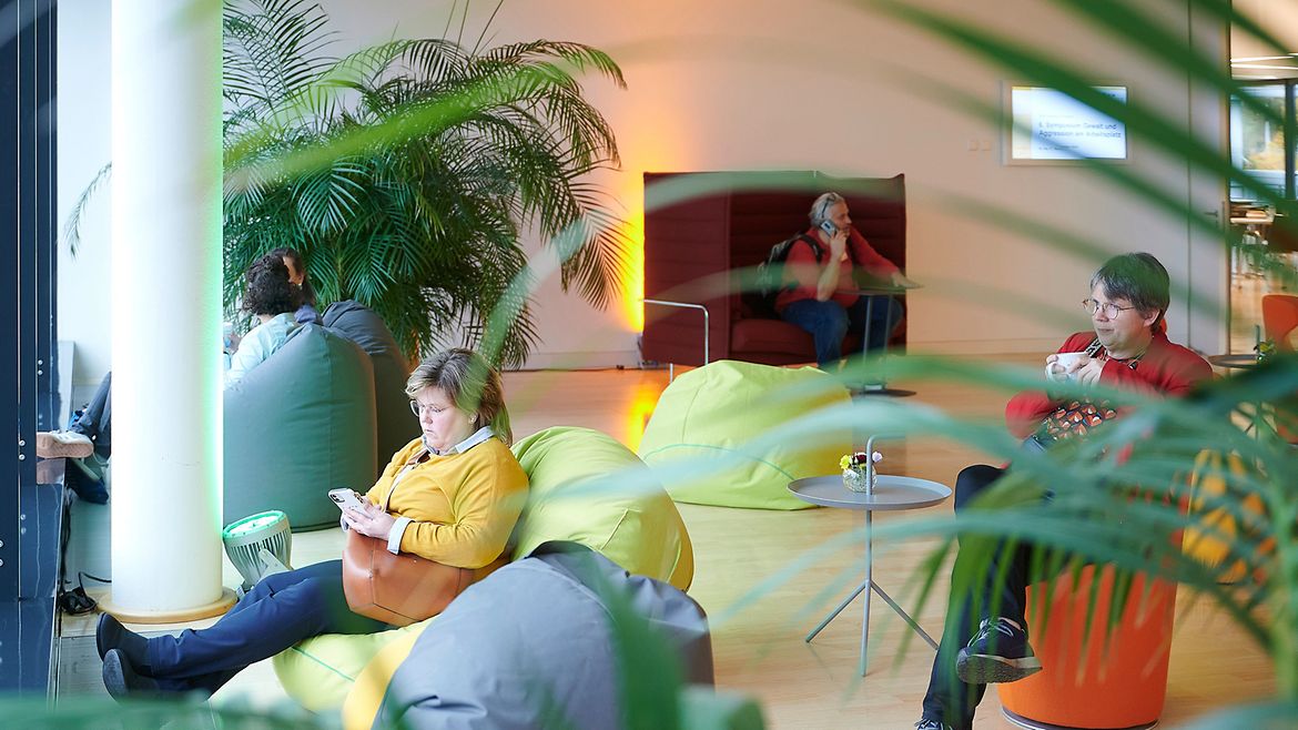Mehrere Menschen sitzen in einem großen Raum verteilt auf verschiedenen Sitzmöbeln und trinken, telefonieren oder schauen aufs Smartphone