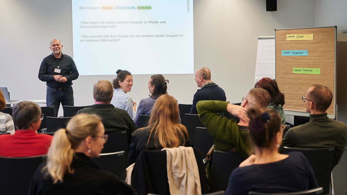 Ein Dozent steht vor sitzenden Teilnehmern einer Veranstaltung, auf der an die Wand projizierten Folie steht "Übung: Vorwurf und Wunsch"