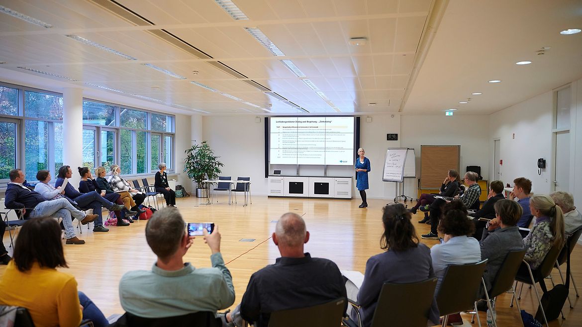 Eine Dozentin steht vor einer Leinwand in einem großen Saal, die Teilnehmerinnen und Teilnehmer sitzen in einem Halbkreis, in der Mitte liegt ein langes rotes Seil