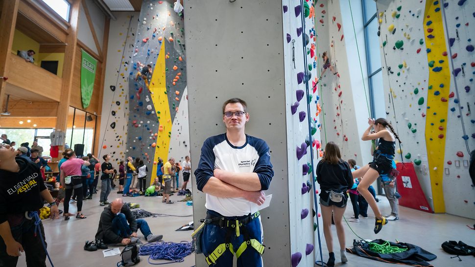 Ein Athlet lehnt an einer Säule in einer Kletterhalle. Im Hintergrund weitere Menschen, teils an Kletterwänden gesichert mit Seilen.