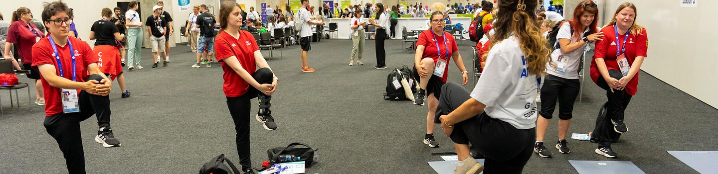 Special Olympics - Healthy Athletes Vier Athletinnen in einer Halle zusammen mit einer Trainerin, die Übungen vormacht. Alle stehen vor einer Yoga-Matte und haben ein Bein zur Brust hochgezogen. Im Hintergrund weitere Menschen.