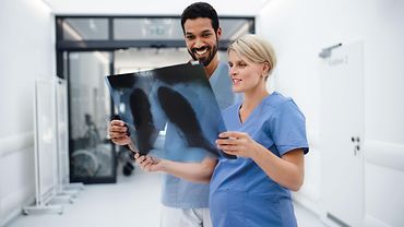 Mann und schwangere Frau in Krankenhaus-Kleidung schauen zusammen auf das Röntgenbild einer Lunge. 