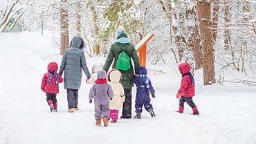 Blick von hinten: Zwei Erwachsene und sechs Kinder laufen in Winterkleidung durch einen verschneiten Wald