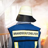 Brandschutz Blick von hinten auf einen Brandschutzhelfer in Warnweste. Er steht auf einer Treppe und schaut von oben auf ein Feuer.