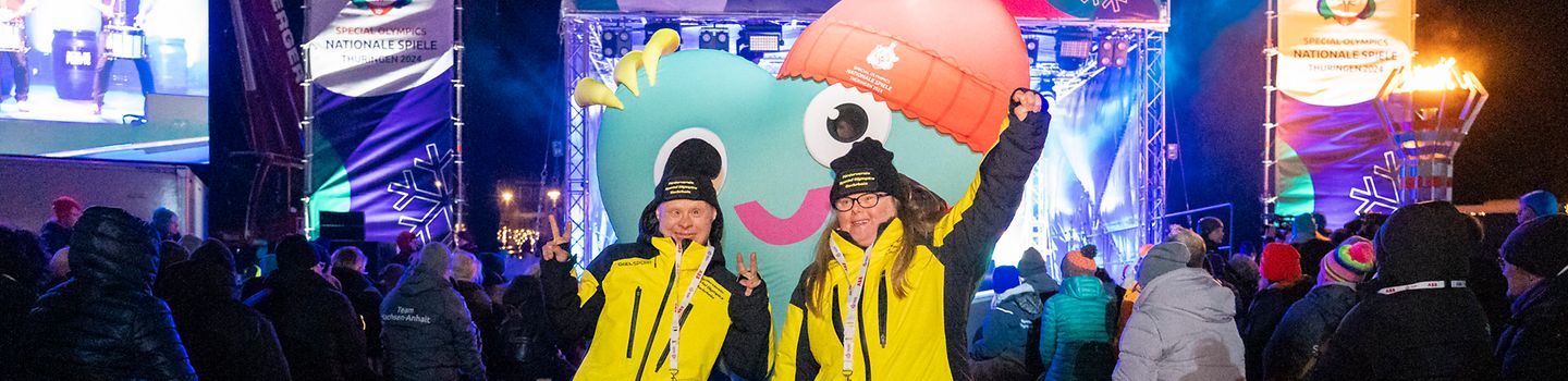 Zwei Teilnehmende tanzen mit dem Maskottchen der Spiele, im Hintergrund die Showbühne und daneben viele Zuschauende.