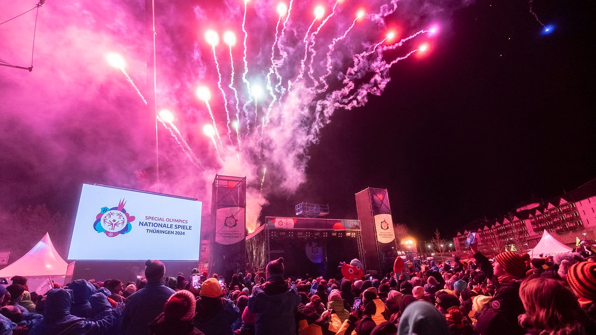 Ein Feuerwerk im Himmel hinter einer Bühne, davor eine Menschenmenge und eine Leinwand mit dem Logo der Spiele