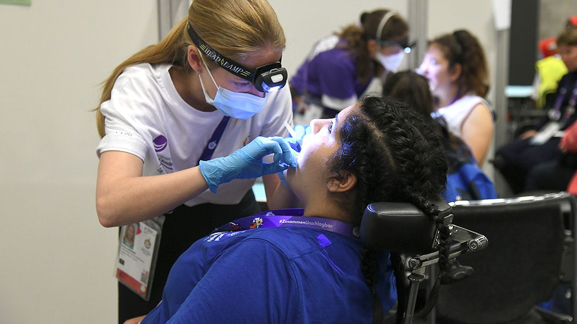Eine Zahnärztin mit Mundschutz, Handschuhen und Stirnlampe leuchtet einer Teilnehmerin in den geöffneten Mund.