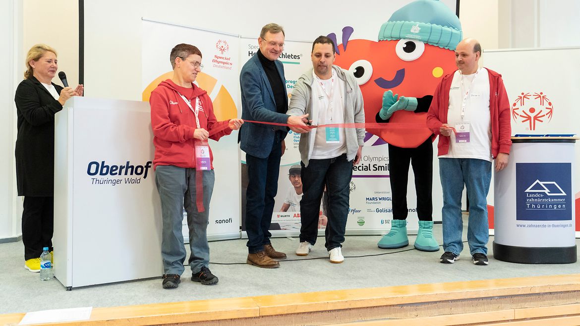 5 Menschen auf einem Podium. Eine Dame links vor einem Mikro. 2 Männer halten ein rotes Band. Der 2. von rechts schneidet es mit einer Schere durch. Im Hintergrund das Herz-Maskottchen der Spiele.