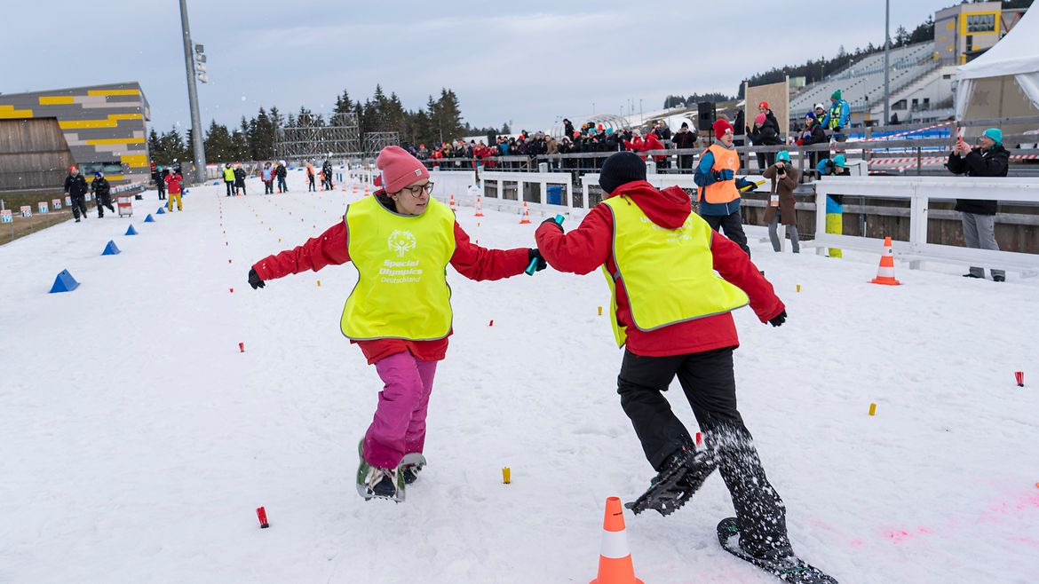 wei Personen in leuchtend gelben Westen und Winterkleidung, die mit Schneeschuhen in einer schneebedeckten Arena laufen. Sie übergeben sich ein Staffelholz. Im Hintergrund sind weitere Teilnehmer und Zuschauer sichtbar, die sich entlang eines abgegrenzten Bereichs aufhalten. 