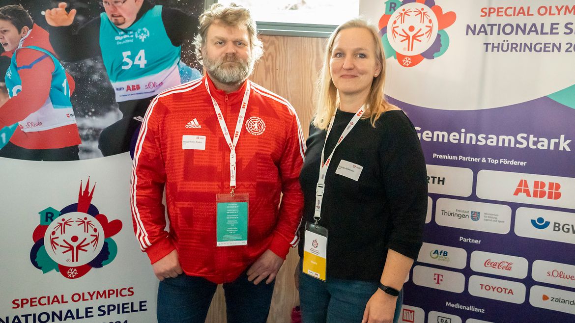 Ein älterer Mann in roter Adidas-Jacke und eine blonde Frau stehen vor 2 Aufstellern mit dem Logo der Spiele und verschiedenen anderen Logos.