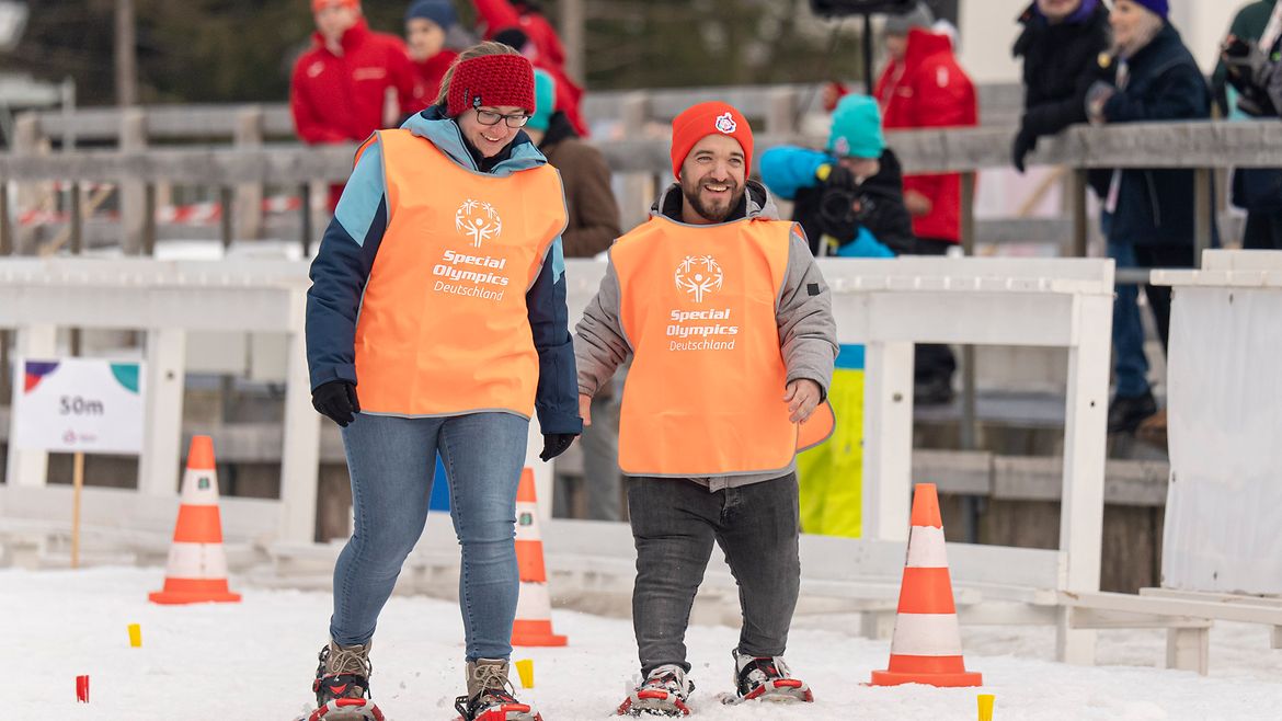 Eine Frau und ein Mann mit Schneeschuhen und orangenen Leibchen mit dem SO-Logo auf einer Schneefläche in einer Arema. Im Hintergrund Zuschauende.