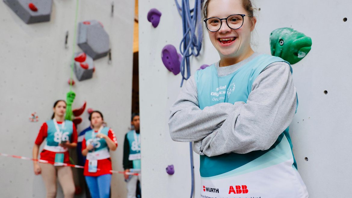 Ein Mädchen mit Brille lehnt an einer Kletterwand und lächelt in die Kamera. Im Hintergrund 2 weitere Kletterinnen.