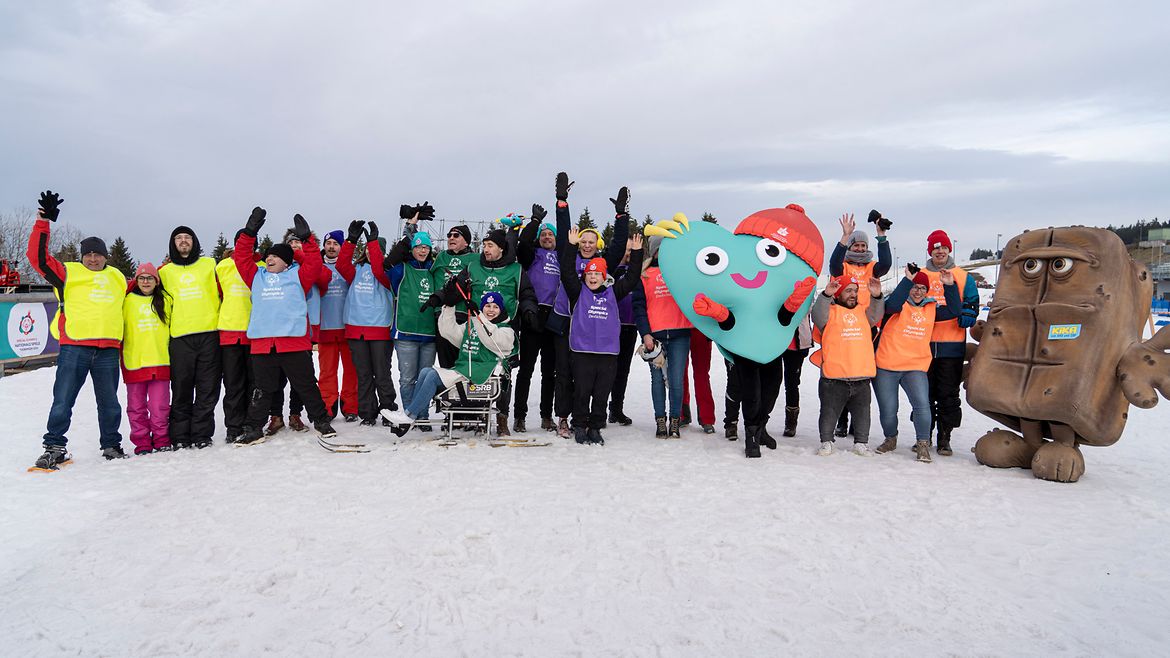 Eine Gruppe von Menschen, die auf einem verschneiten Feld stehen und ihre Hände in die Luft heben. Zwei große Maskottchen sind ebenfalls zu sehen. Die Menschen tragen verschiedene farbige Westen, darunter Gelb, Blau, Grün und Rot. Eines der Maskottchen sieht aus wie ein fröhliches, buntes Herz, während das andere wie ein braunes Brot mit einem Gesicht darauf aussieht.