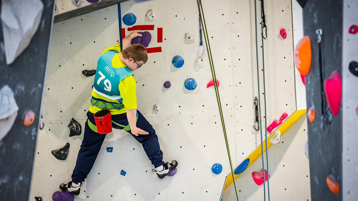 Ein Junge klettert an einer Kletterwand. Er ist oben angekommen und macht mit der Hand ein Zeichen Richtung Boden.