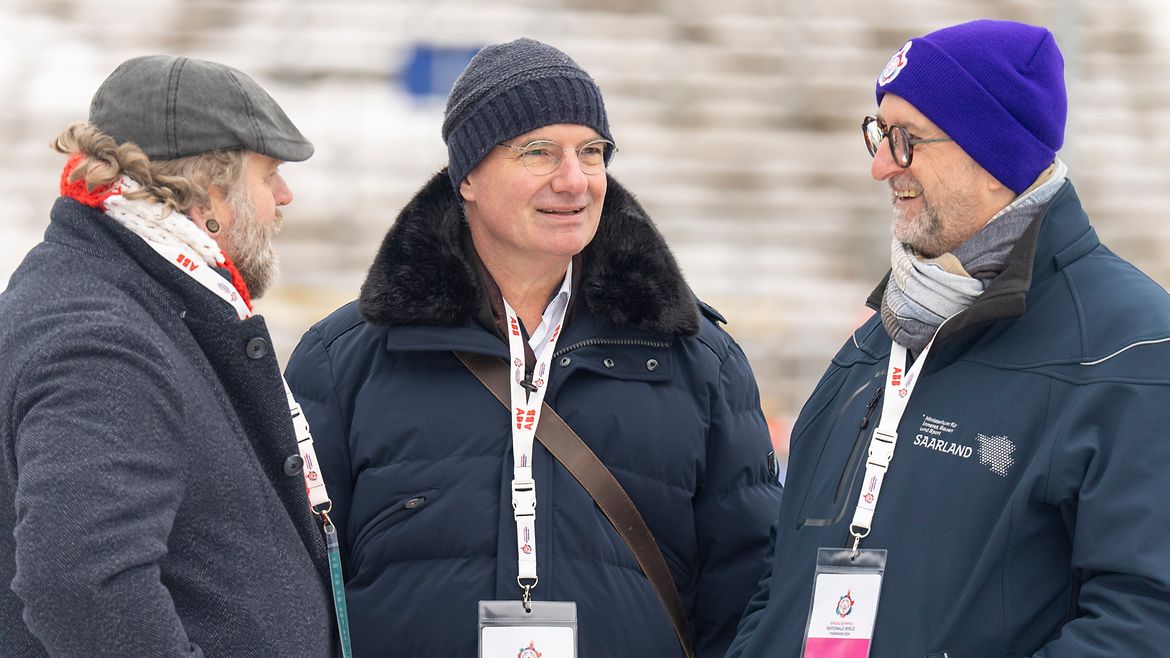 Drei Männer im Gepräch. Alle tragen Mützen, zwei eine Brille. Im Hintergrund Treppen eines Stadions.